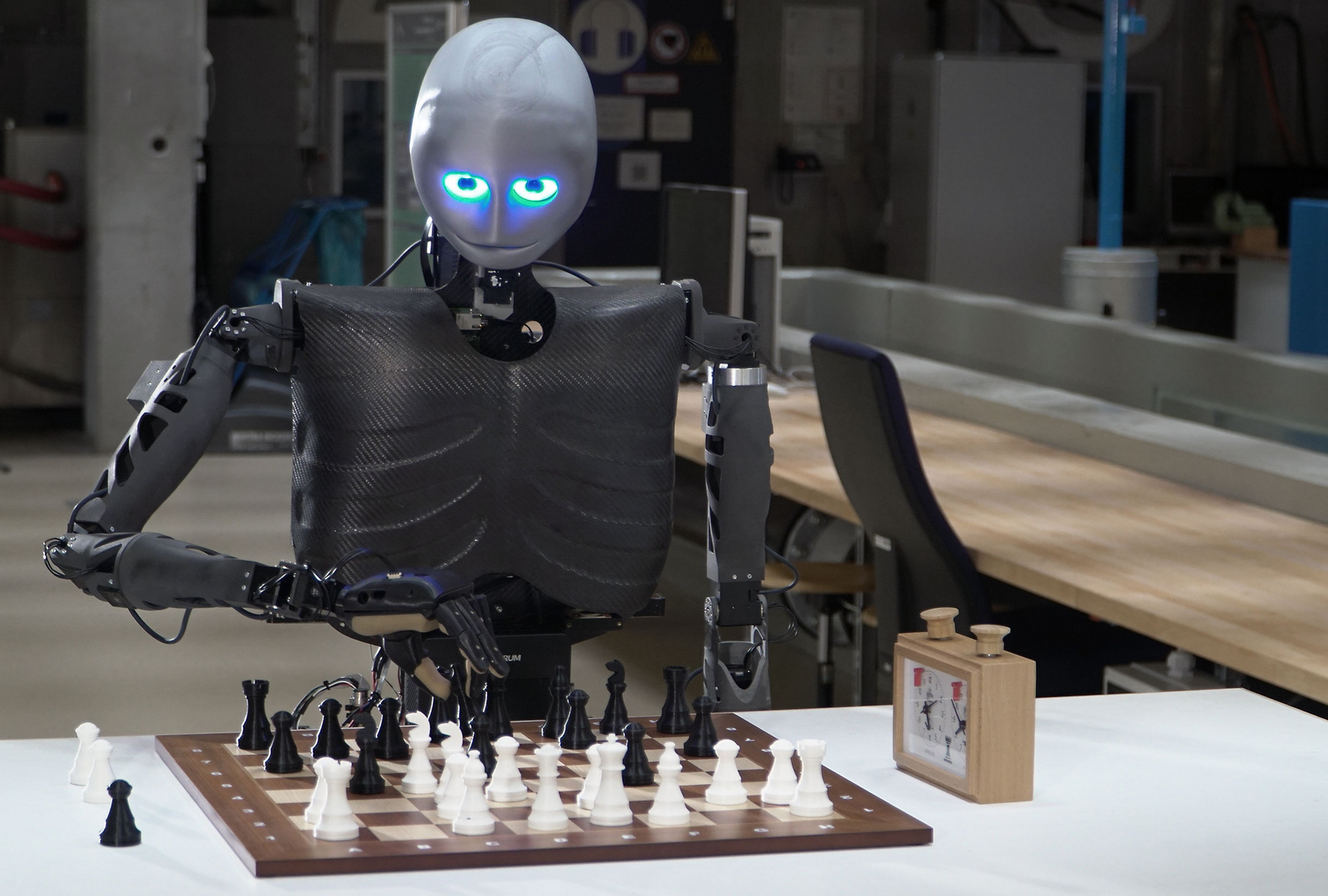 Der humanoide Roboter Sweaty sitzt an einem Tisch, vor sich ein Schachbrett mit Figuren, rechts davon eine Schachuhr