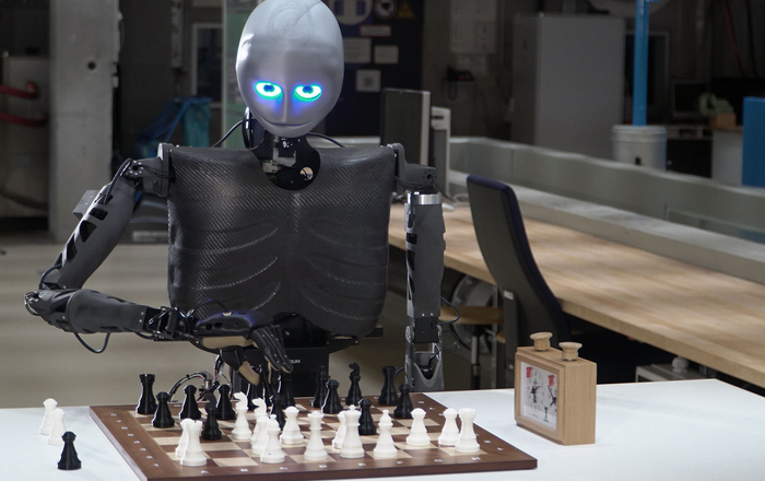 Der humanoide Roboter Sweaty sitzt an einem Tisch, vor sich ein Schachbrett mit Figuren, rechts davon eine Schachuhr