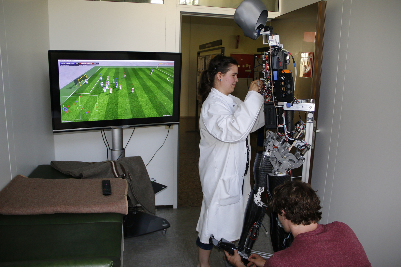 Zwei Studierende arbeiten im Sanitätsraum der Hochschule Offenburg daran, Sweaty wieder fit zu machen während dieser auf einen Bildschirm schaut auf dem ein Spiel des Teams Magma läuft.