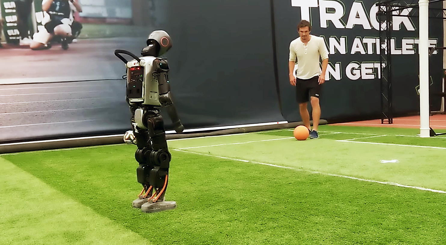 Ein Mann spielt einem humanoiden Roboter auf einem Fußballfeld einen Ball zu.
